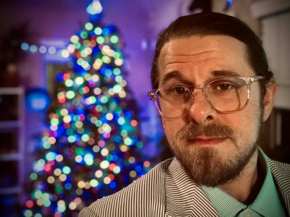 A self-portrait of Jared Caraway standing in front of a brightly lit Christmas tree.
